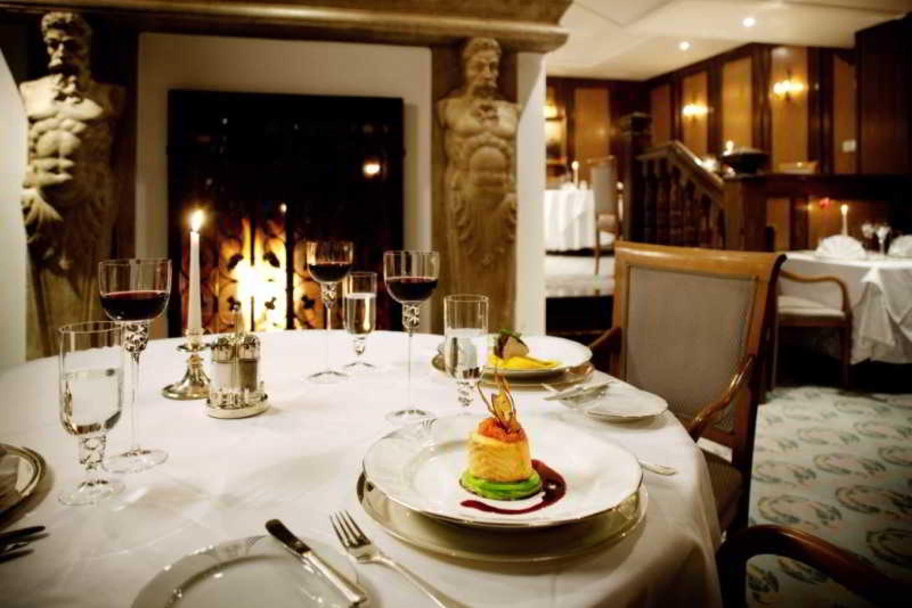 Art Nouveau Palace Hotel Praha Restoran foto The photo depicts an elegant dining setting in a restaurant. In the foreground, there is a round table elegantly laid with fine dining utensils, including plates, cutlery, and glasses. 

On the table, a beautifully presented dessert sits on a plate, 