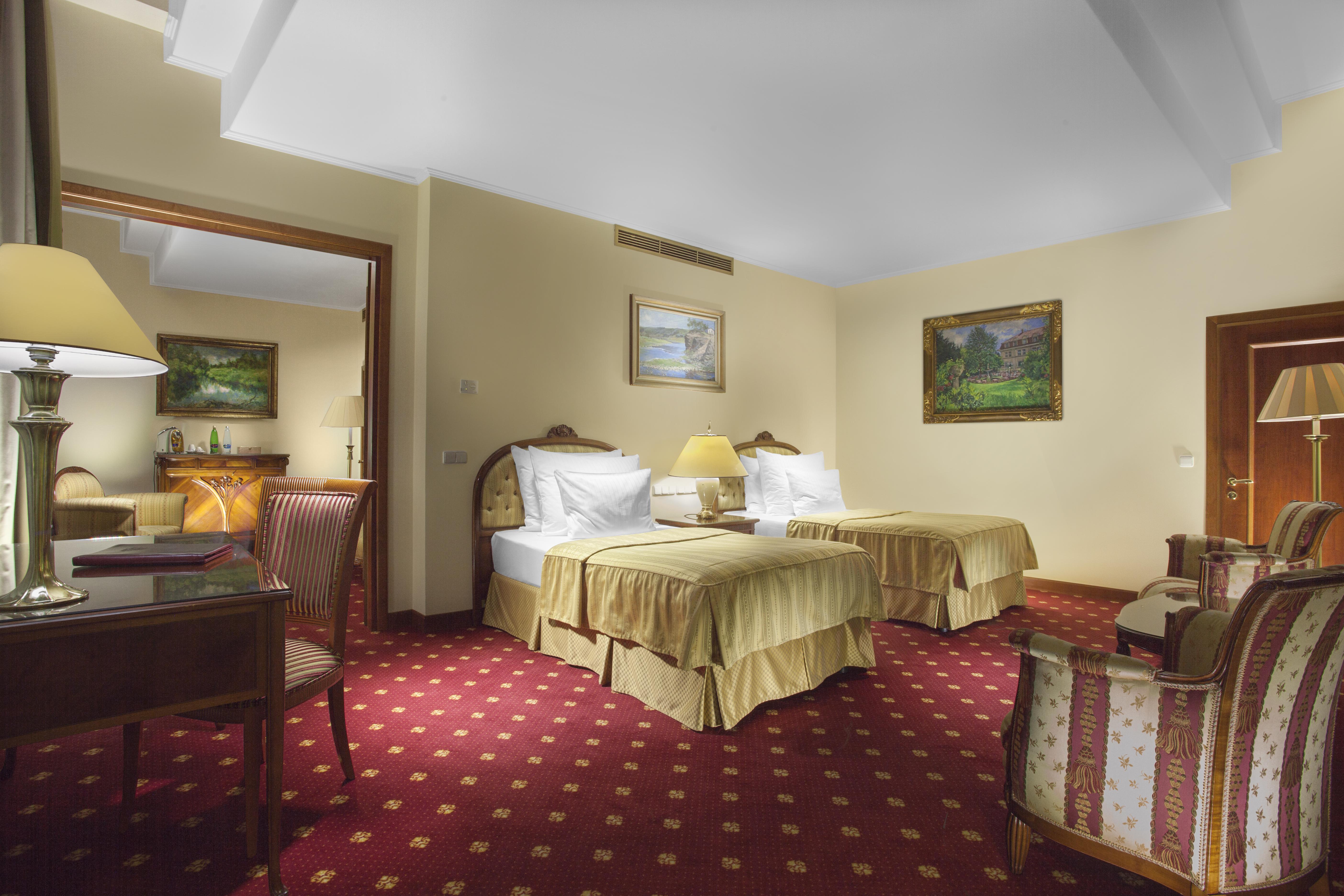 Art Nouveau Palace Hotel Praha Bagian luar foto The photo depicts a hotel room featuring two beds with soft, golden bedspreads. The room is furnished with a small desk and a chair, along with a lamp on the desk. There are decorative paintings on the walls and a plush carpet with a red pattern. The