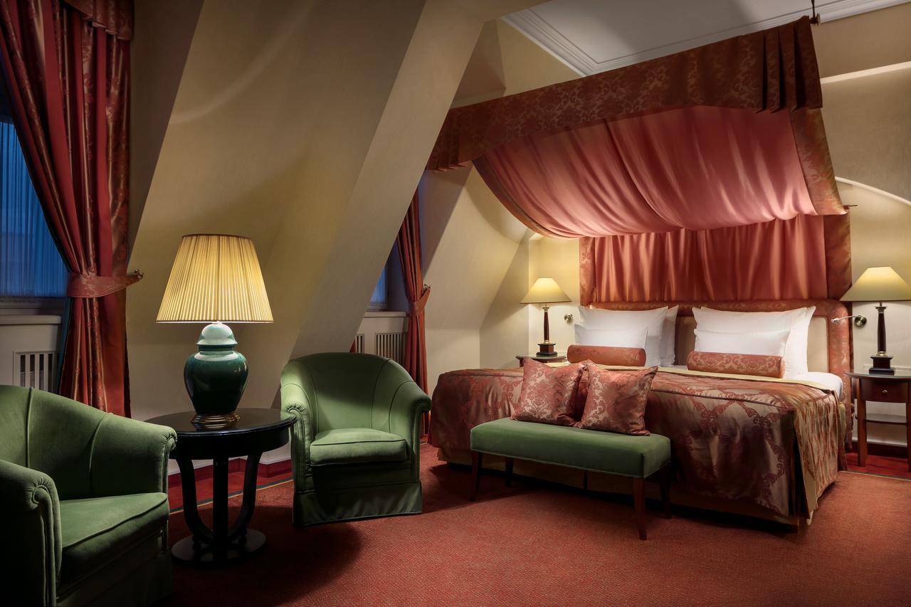 Art Nouveau Palace Hotel Praha Bagian luar foto The photo depicts a cozy and elegantly furnished hotel room. It features a bed with a canopy draped in pink fabric, complemented by rich red and gold decor. There are two green armchairs and a small round table near the bed, creating a relaxed seatin